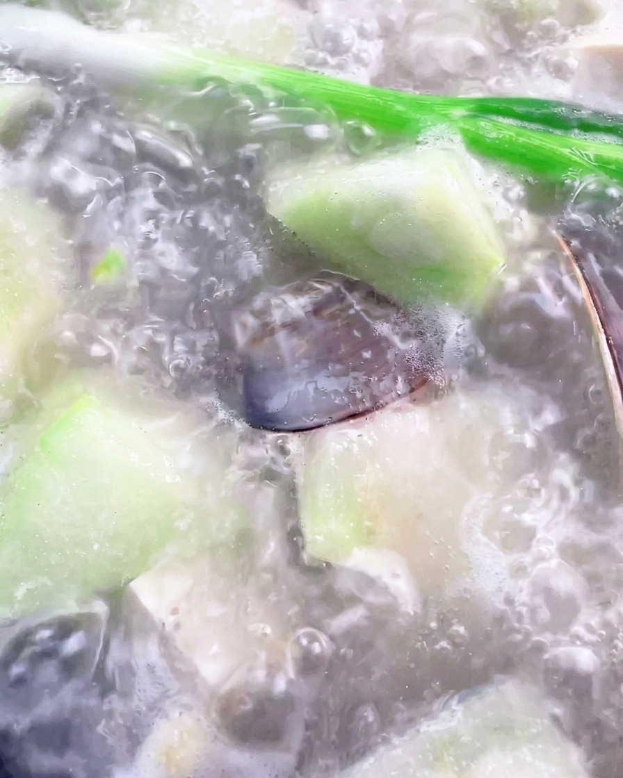 A ladle removing white foam from the surface of a boiling luffa and clam soup in a pot.