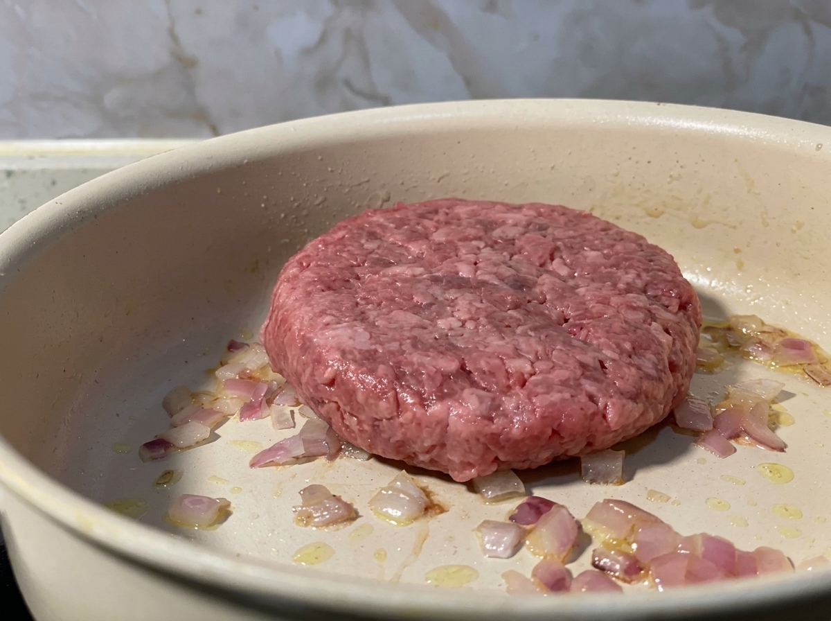 A raw ground beef patty placed in a pan alongside sautéed red onions.