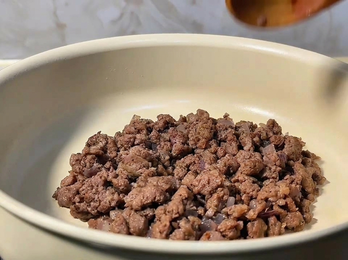 Ground beef being stirred and broken up in a pan with sautéed onions.