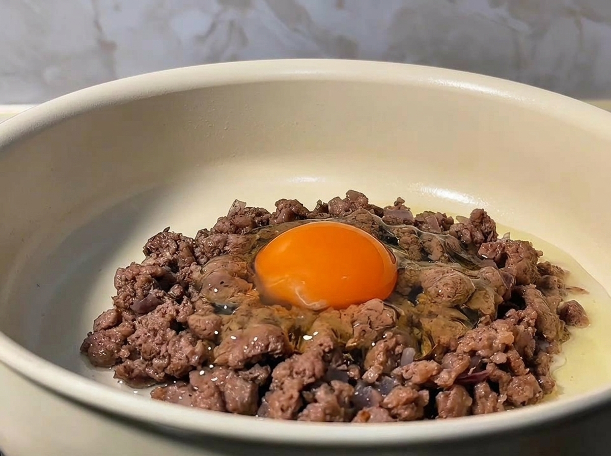 A raw egg cracked into the center of browned ground beef in a pan.