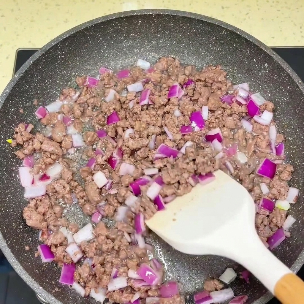 Sautéed ground beef and chopped red onion being seasoned with sauces in a frying pan.