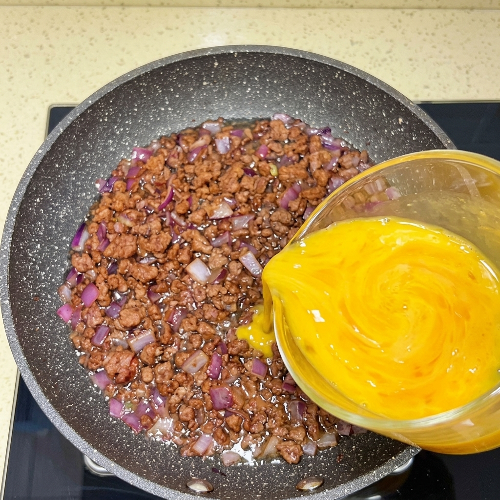 Beaten eggs being poured from a glass measuring cup into a pan with browned beef and onions.