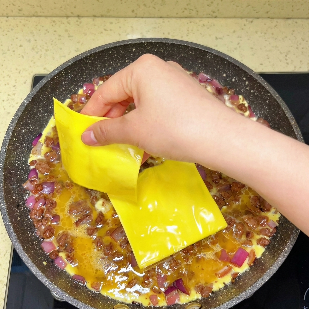 Slices of processed cheese being layered on top of cooking eggs and ground beef in a skillet.
