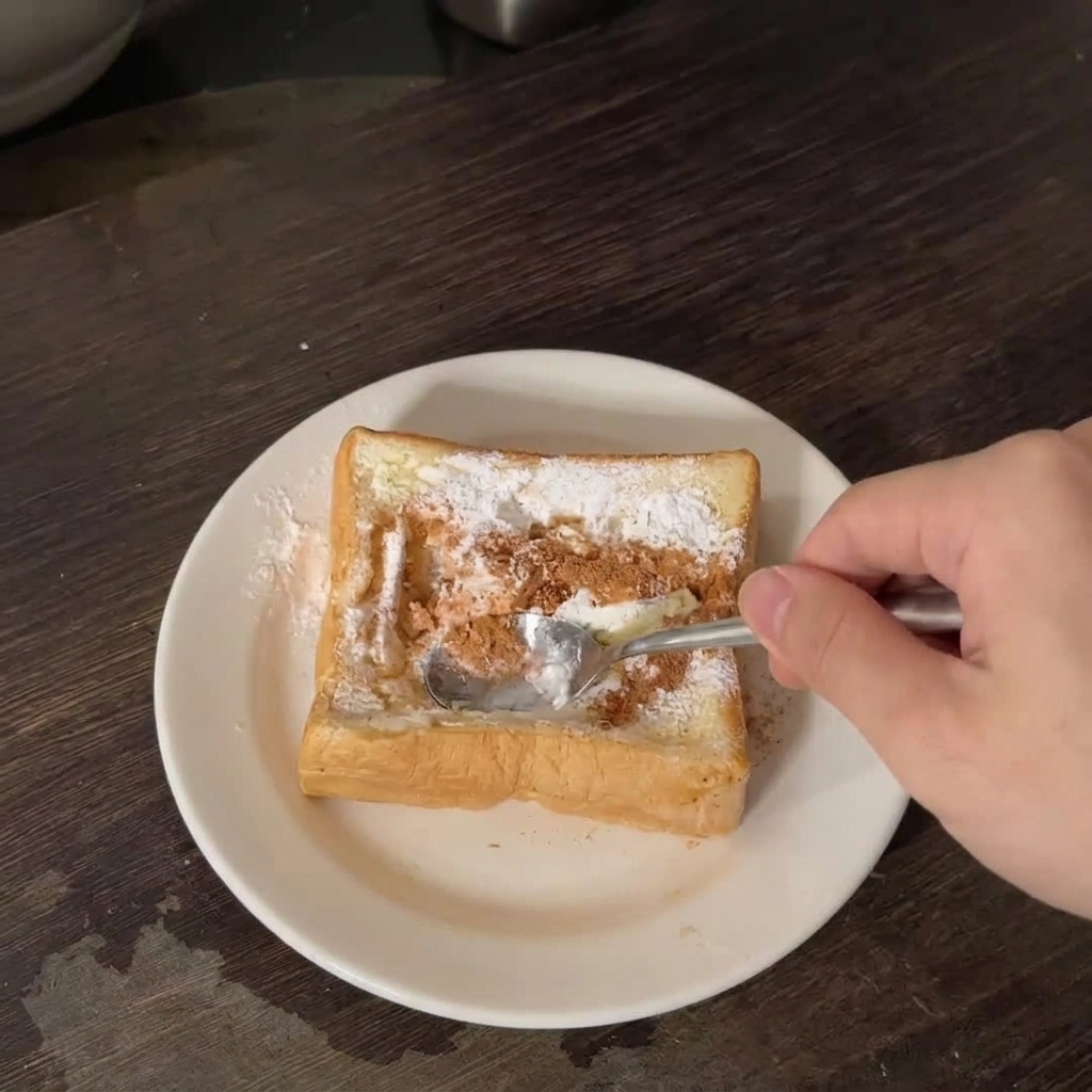 A hand using a metal spoon to press down the center of a thick slice of white toast sprinkled with seasoning on a white plate.