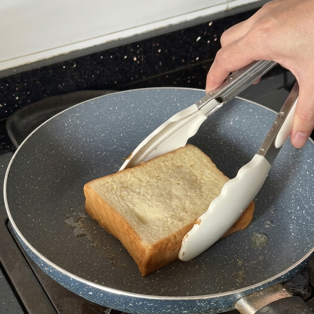 Thick slice of bread being toasted in a frying pan using white tongs.