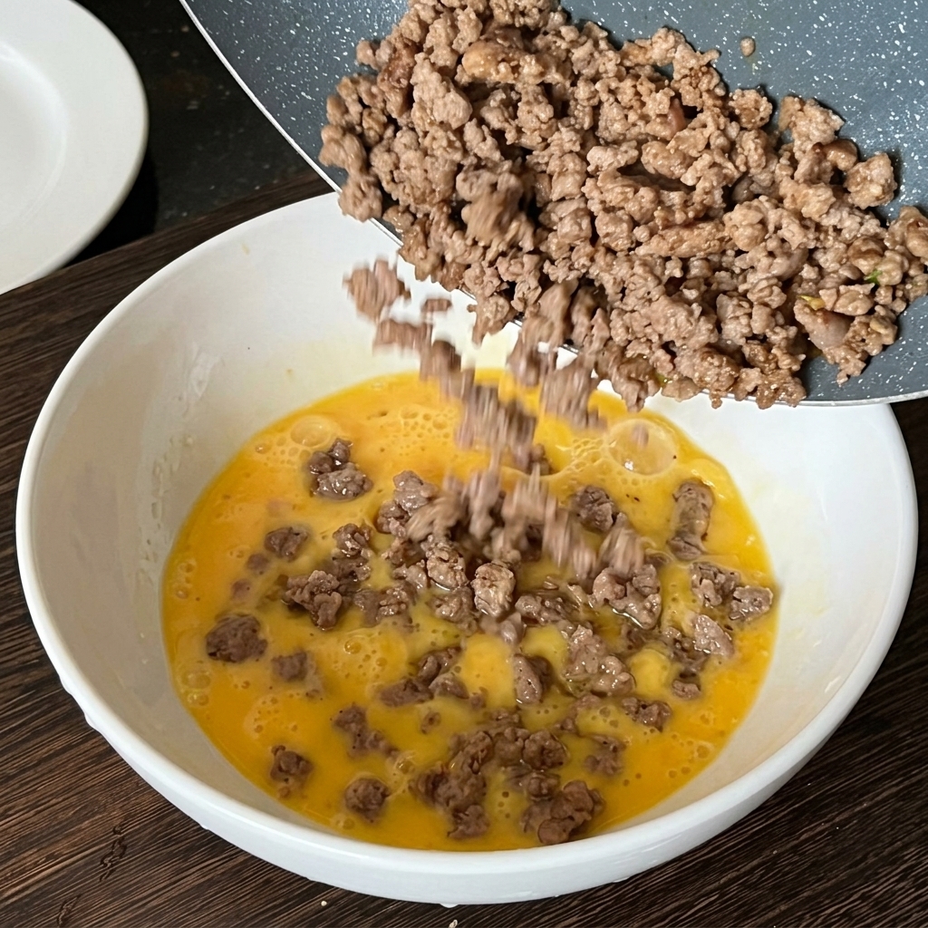 Cooked minced beef being poured from a frying pan into a bowl of raw beaten eggs.
