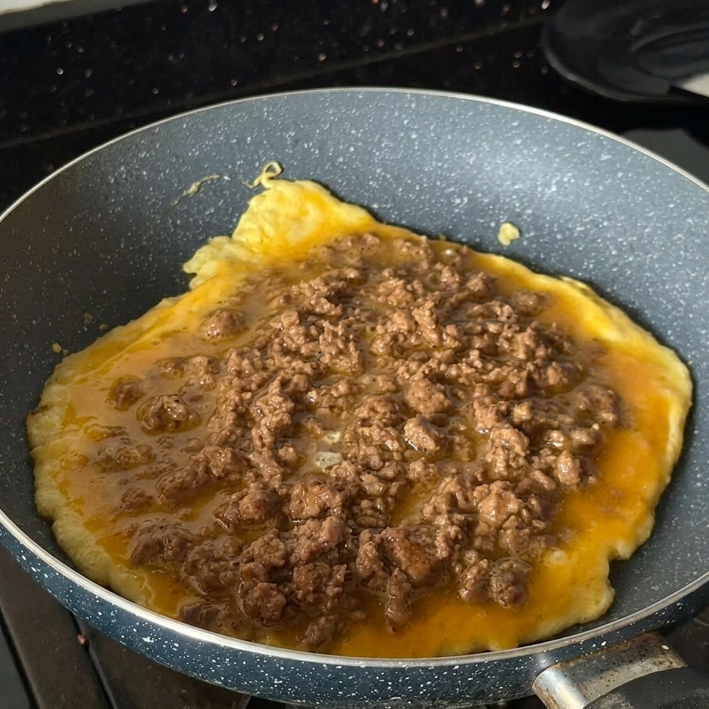 A round egg omelet cooking in a speckled pan, heavily topped with cooked, seasoned minced beef.