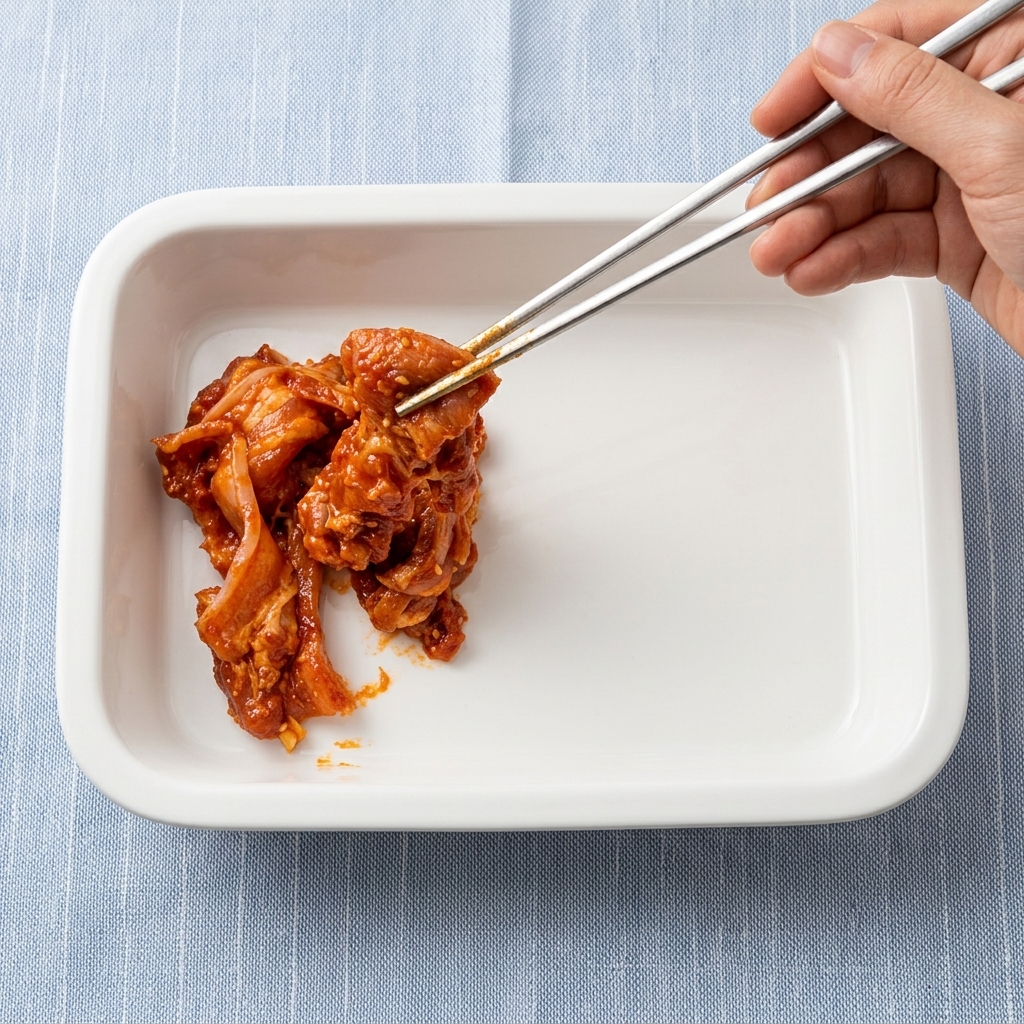Marinated pork belly strips being placed into a white baking dish with metal chopsticks.