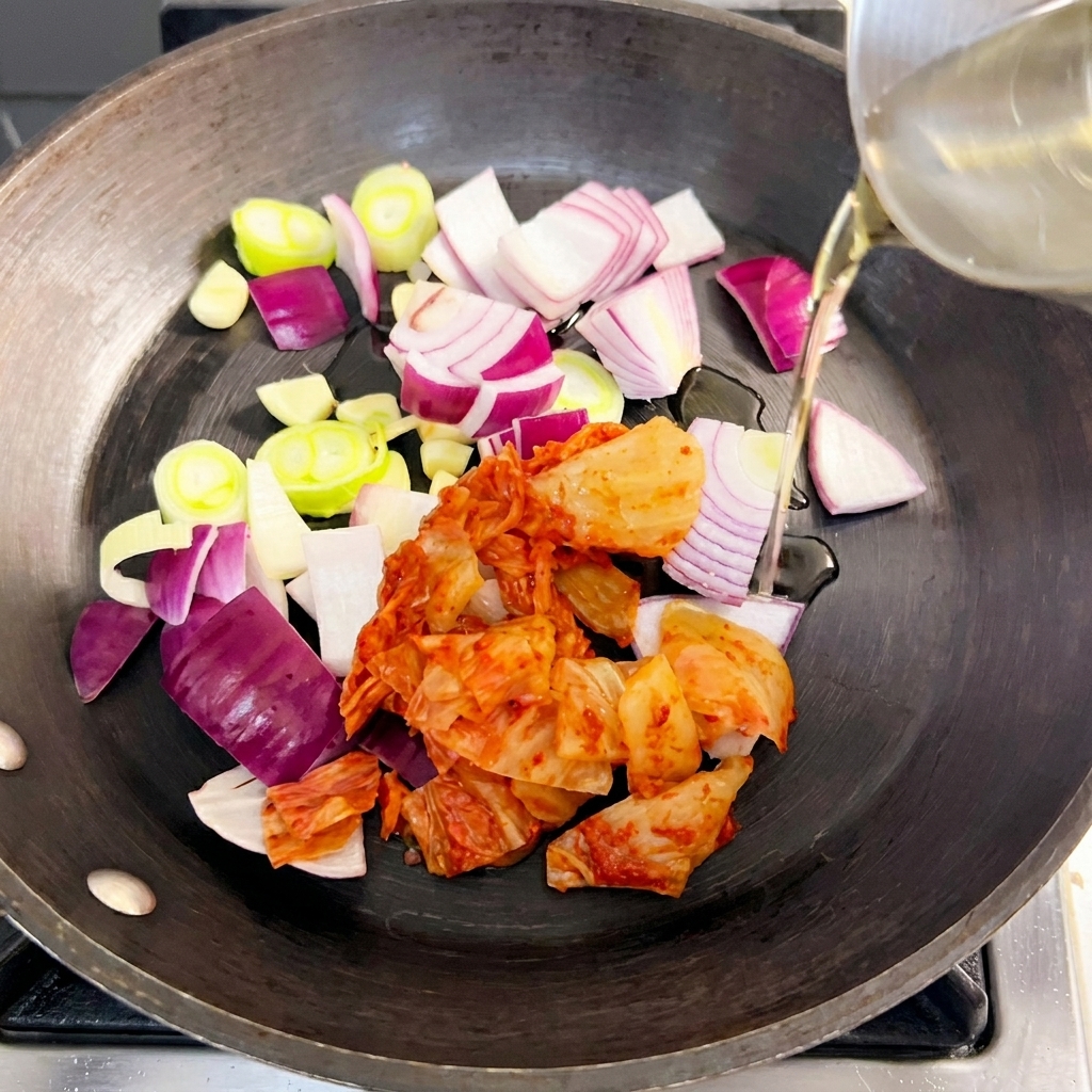 A pan containing sliced red onion, green onion, and kimchi being stir-fried in oil.
