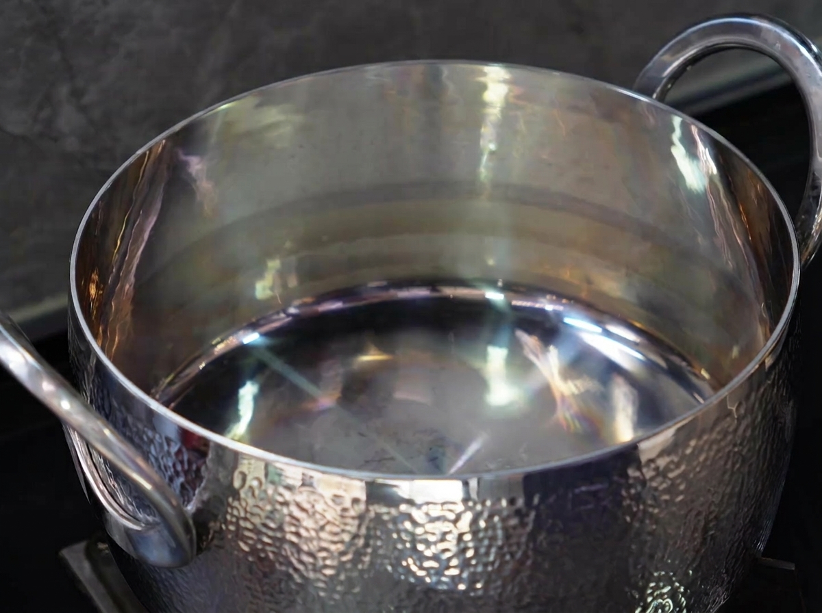A close-up view of a spoonful of clear cooking oil being poured into an empty, shiny stainless steel pot.