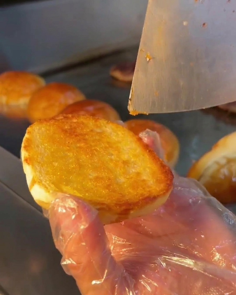 A metal spoon spreading a creamy white sauce onto the toasted surface of a burger bun.