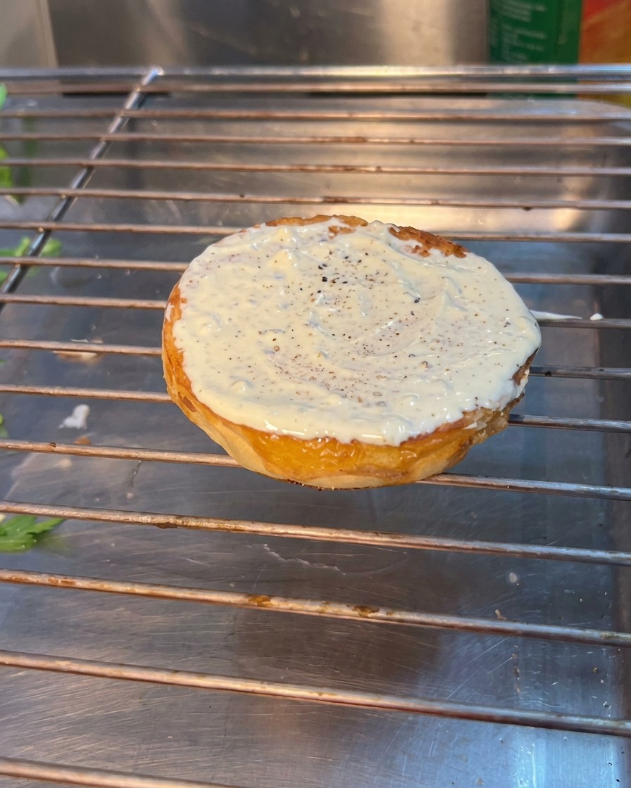 A burger assembly showing arugula, tomato, and pickles layered onto the bottom toasted bun.