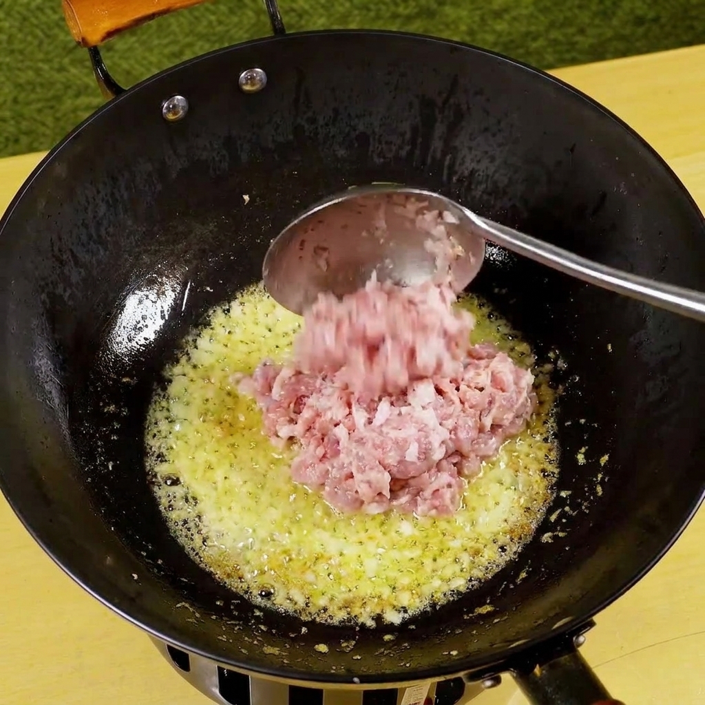 Minced pork being added to a pan with sautéed onion and garlic, beginning to brown.
