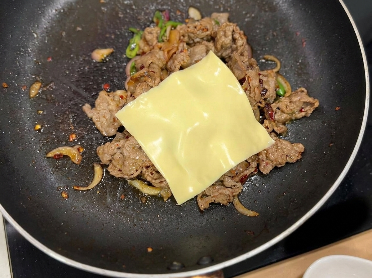 A slice of yellow cheese melting over the cooked beef and onion mixture in the pan.