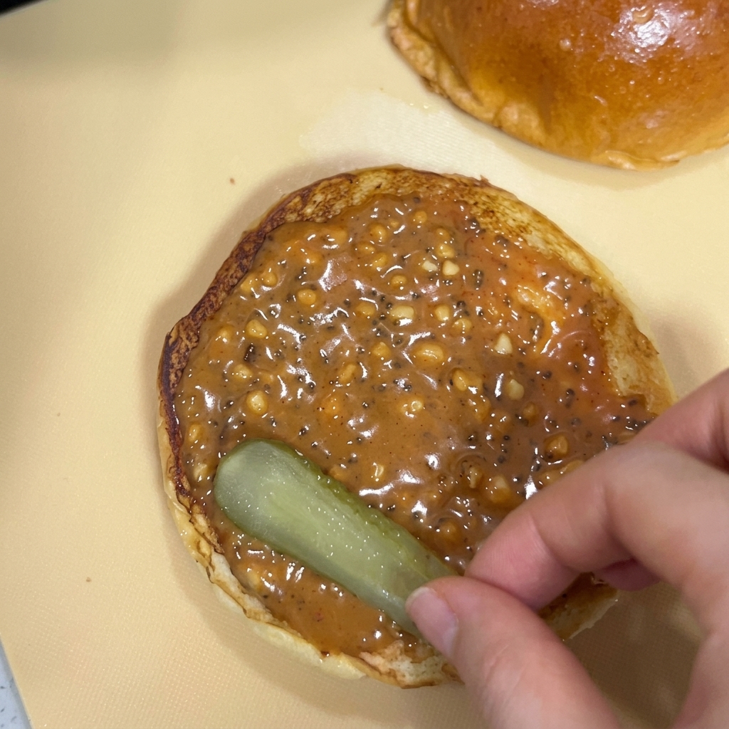 A hand placing a slice of pickled cucumber onto a burger bun covered in thick peanut sauce.