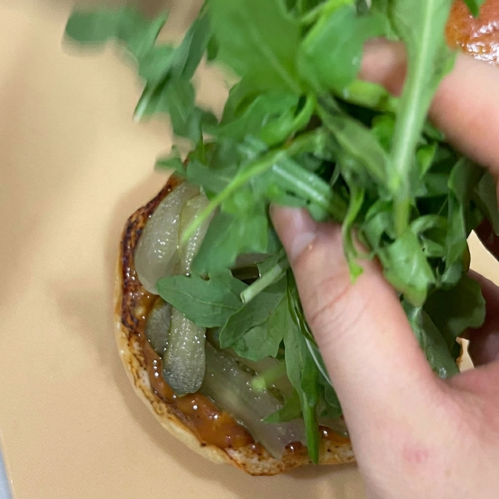 A hand piling fresh green arugula leaves on top of pickles on a burger bun base.