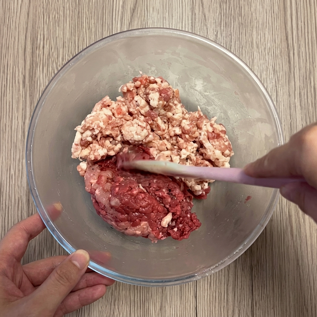Ground pork and beef being combined in a clear mixing bowl.