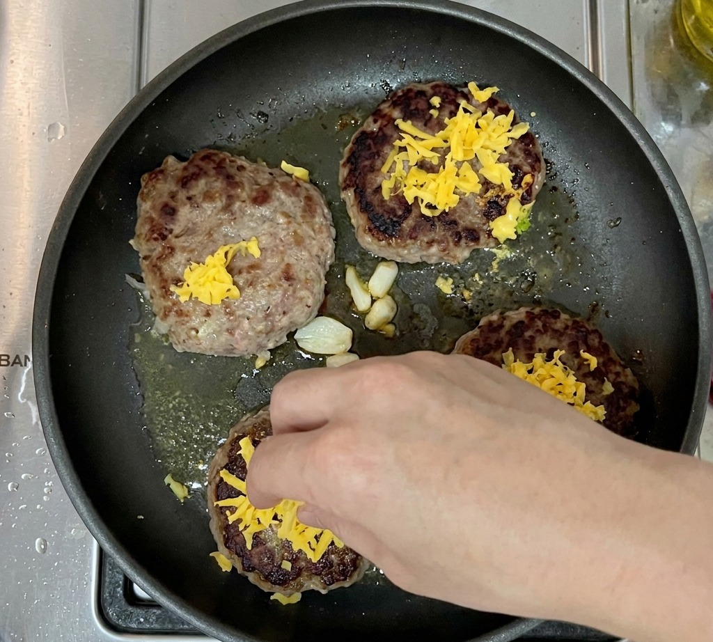 Four cooked burger patties in a pan, each topped with a mound of shredded cheddar cheese.