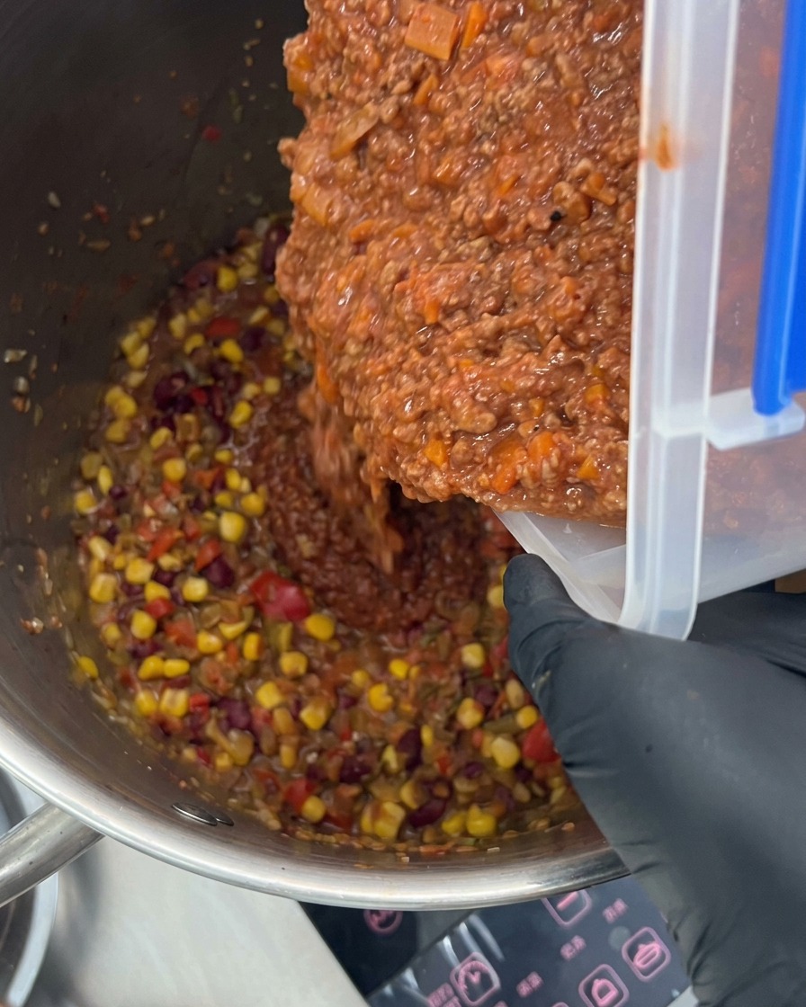 Rich meat sauce being poured into a large stockpot filled with colorful vegetables and spices.