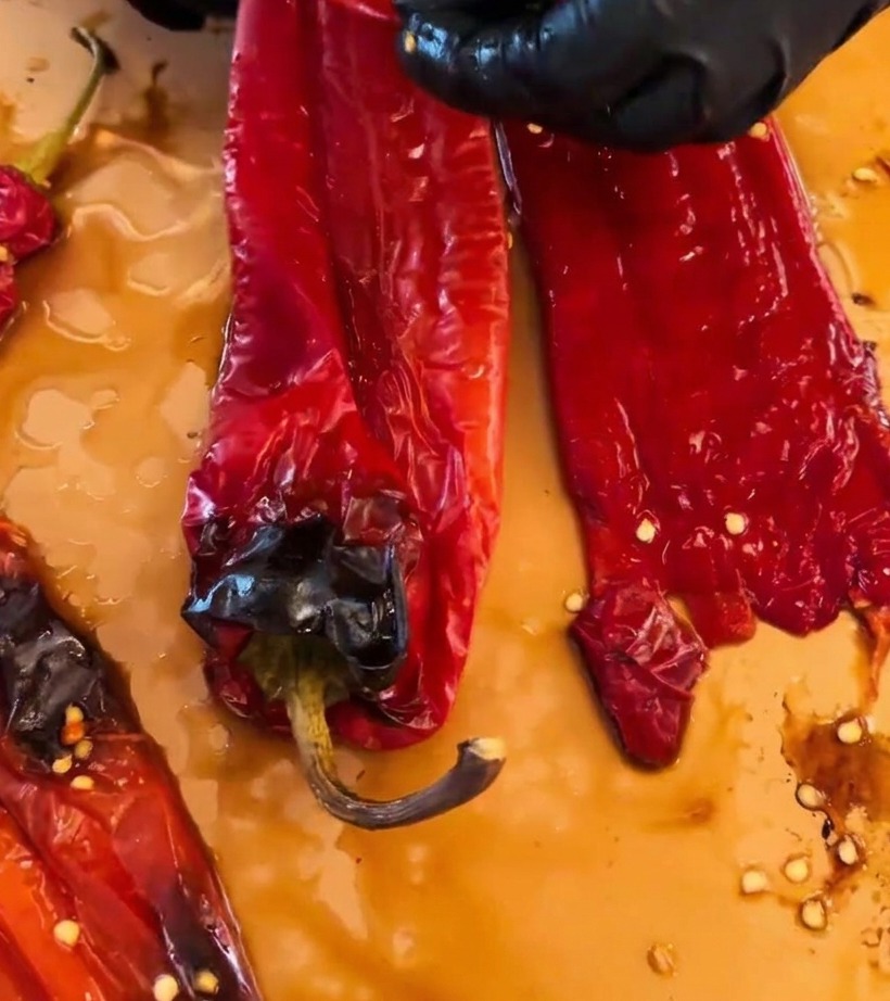 Hands wearing black gloves peeling charred skin off roasted red chilies on a cutting surface.