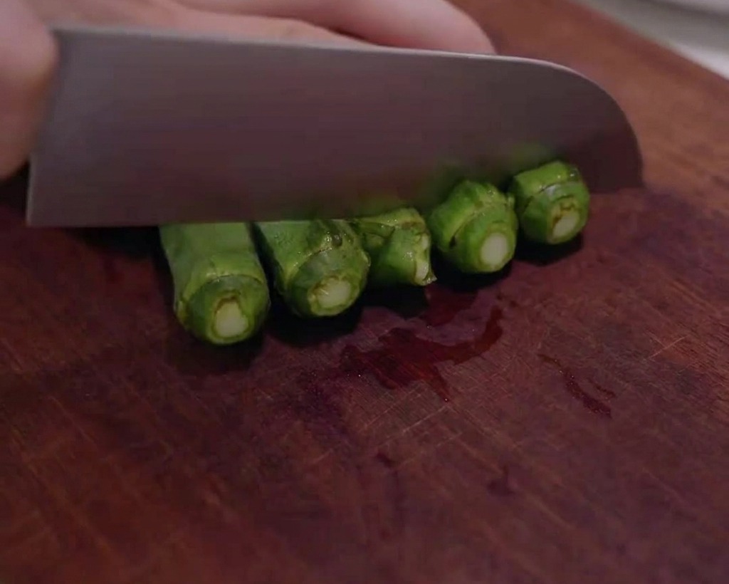 Fresh green okra being sliced into small rounds on a wooden cutting board.