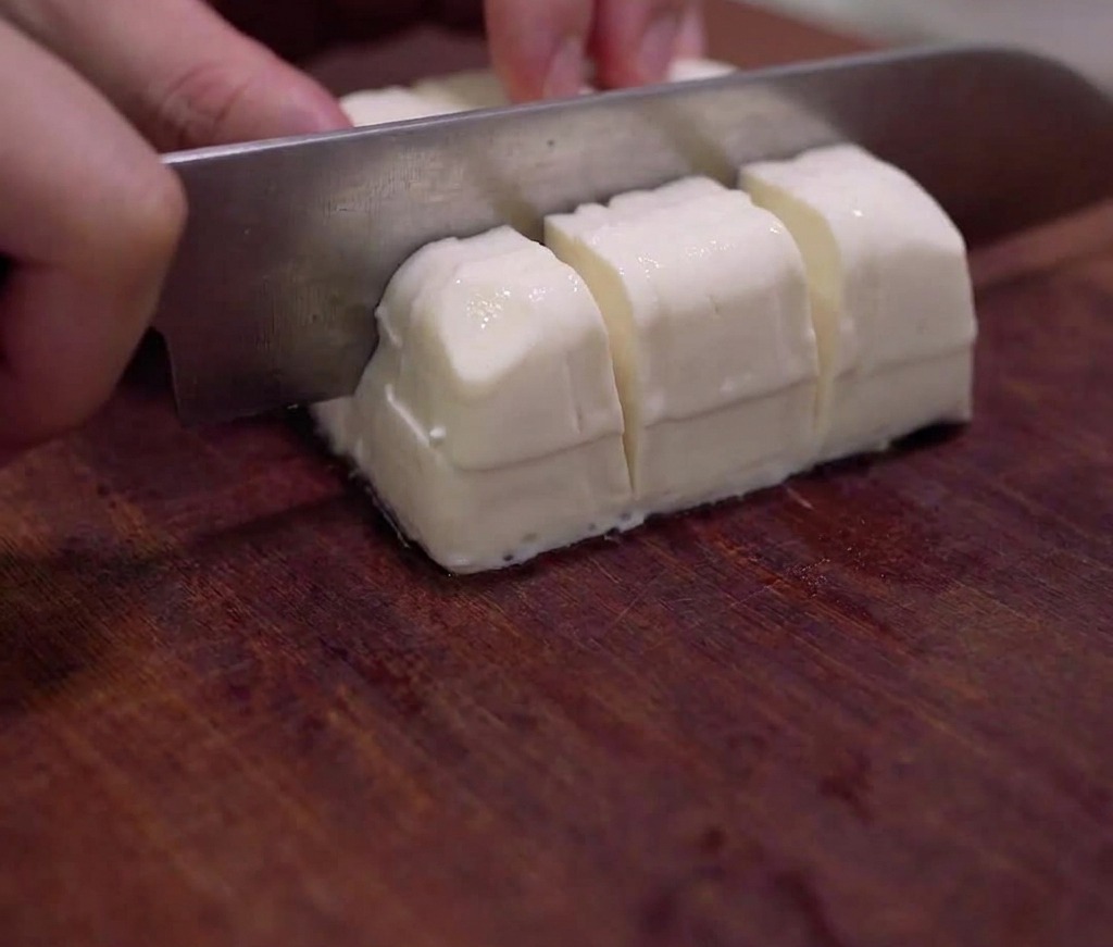 A block of silken tofu being carefully cut into small cubes on a wooden cutting board.