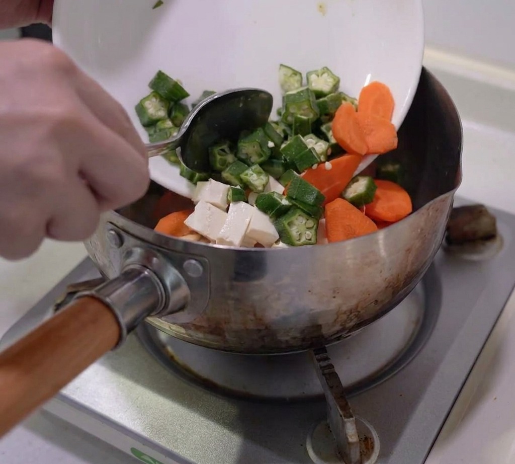 Prepared okra, carrots, mushrooms, and tofu being added to a stainless steel cooking pot.