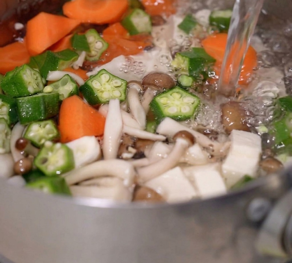 Boiling water being poured into a pot containing mixed vegetables and tofu.