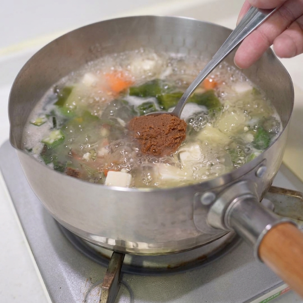 A spoonful of brown miso paste being lowered into the boiling vegetable soup.