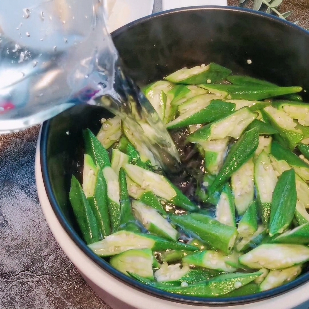 Water being poured into a pot containing stir-fried okra pieces.