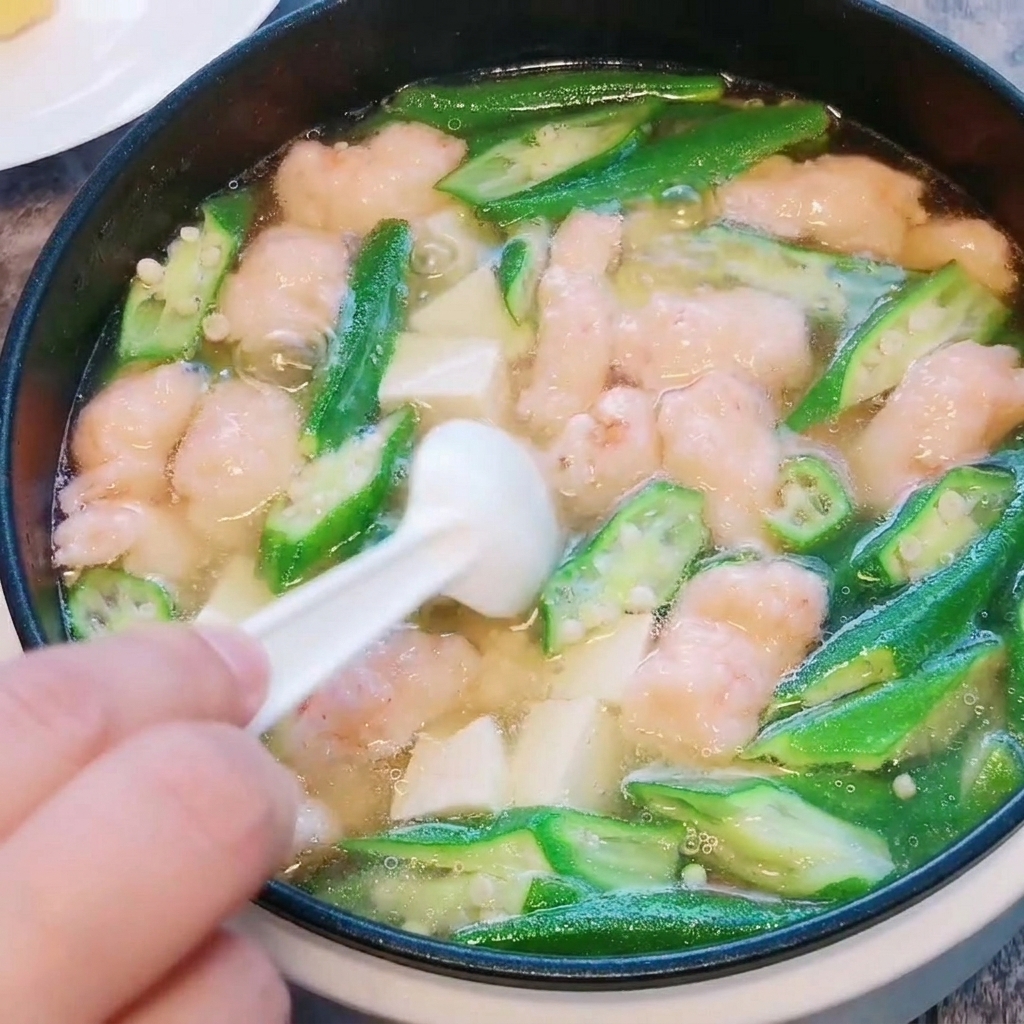 A close-up of salt and white pepper being sprinkled into the finished okra, tofu, and shrimp paste soup.