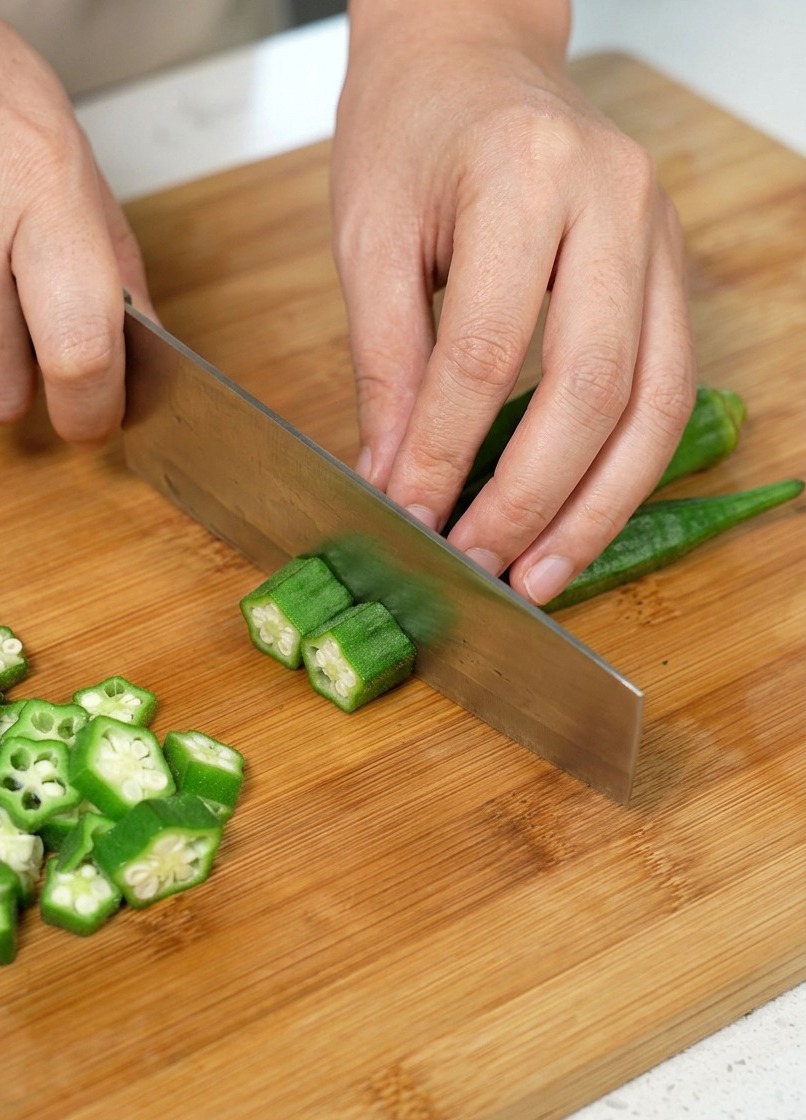 Okra being sliced into rounds on a wooden cutting board.
