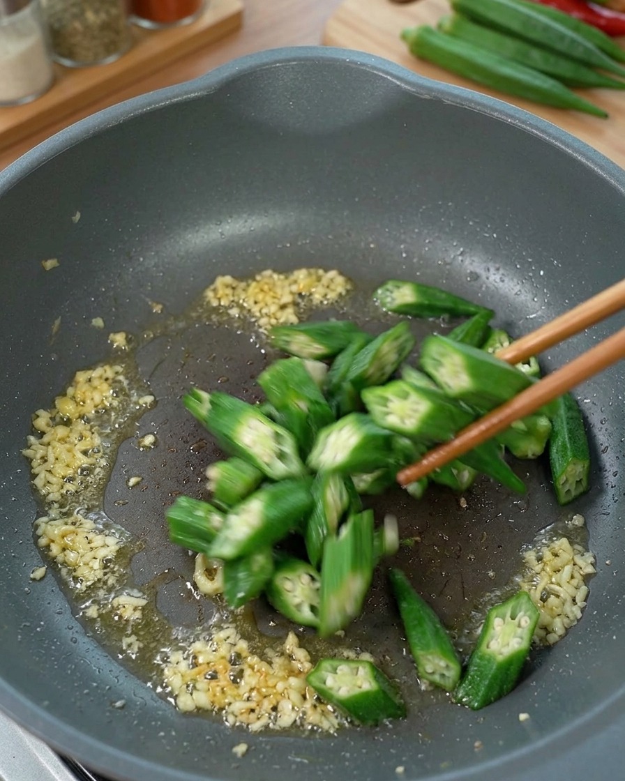Sliced okra being stir-fried in a pan with ginger and oil.