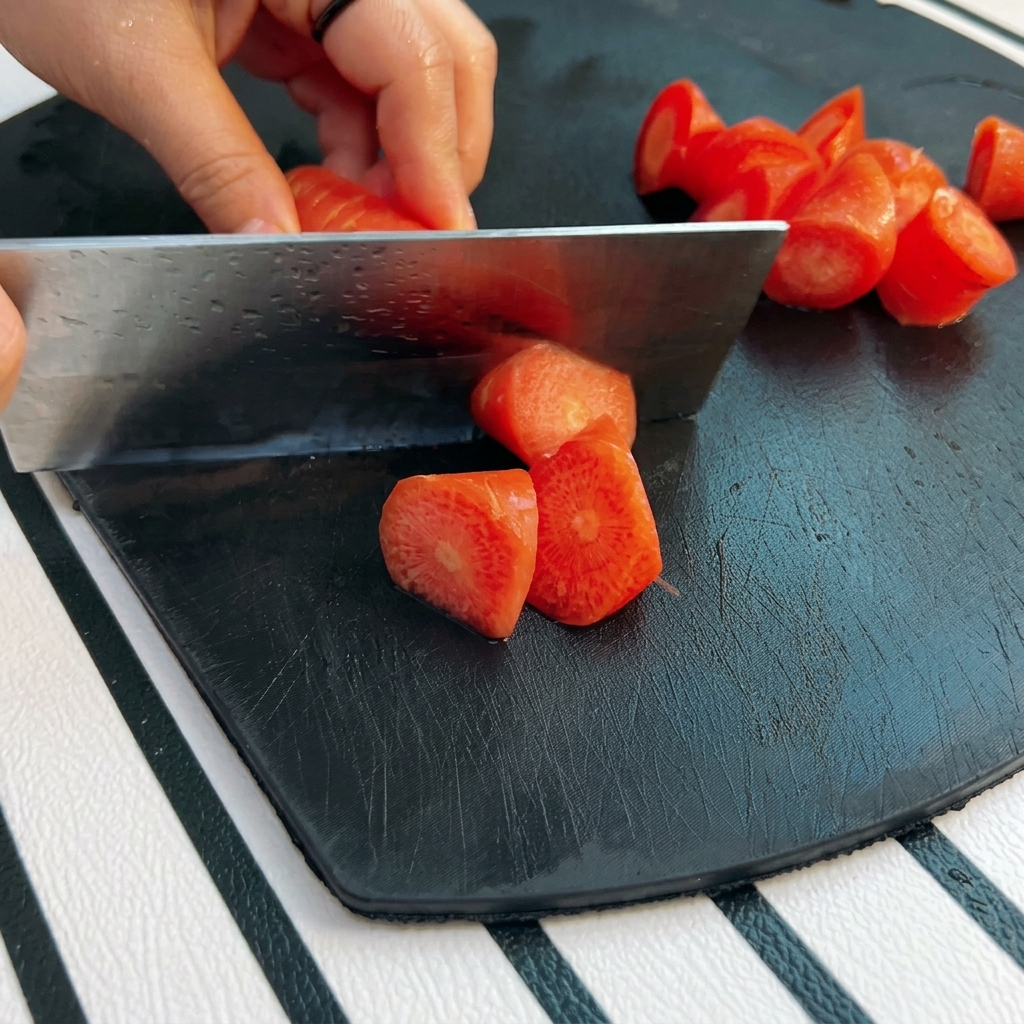 A hand using a large metal cleaver to cut a bright orange carrot into chunky roll-cut pieces on a black board.