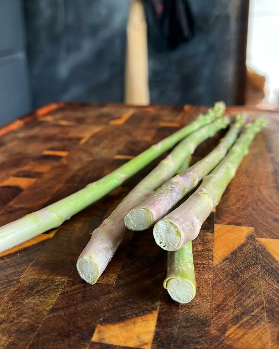 A close-up of fresh green asparagus spears with the tough bottom ends being cut off with a knife on a wooden board.