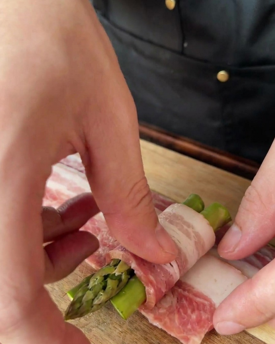Hands wrapping a thin slice of bacon around a bundle of three asparagus spears on a wooden cutting board.