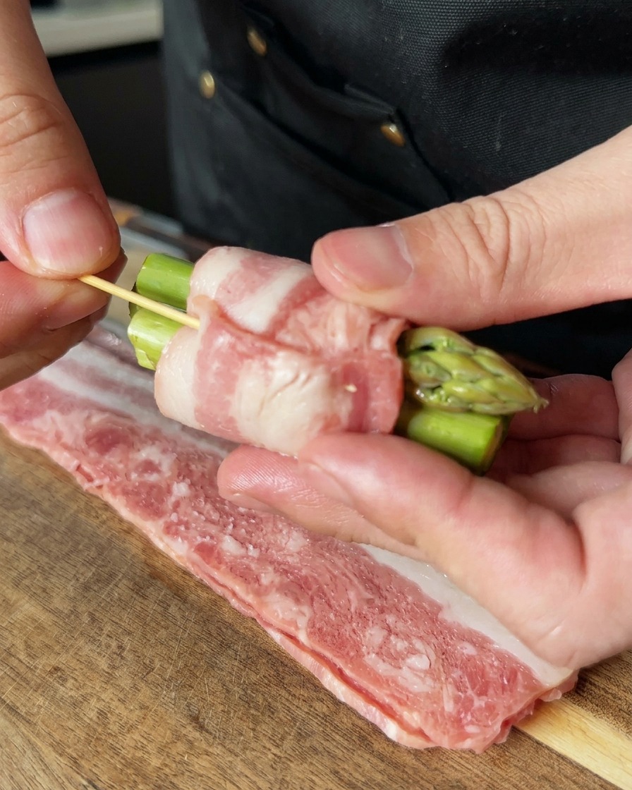 A hand using a wooden toothpick to secure a bacon-wrapped asparagus bundle, holding the ingredients firmly together.