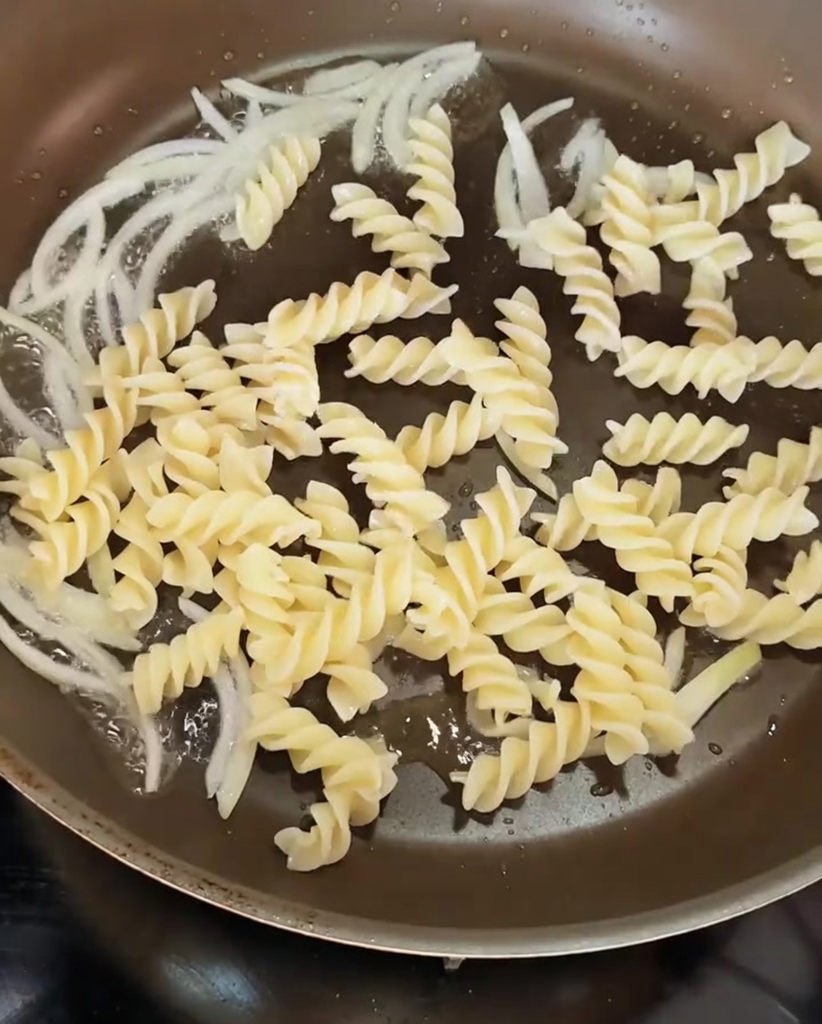 Cooked spiraled pasta being added to a pan containing sautéed onion slices.