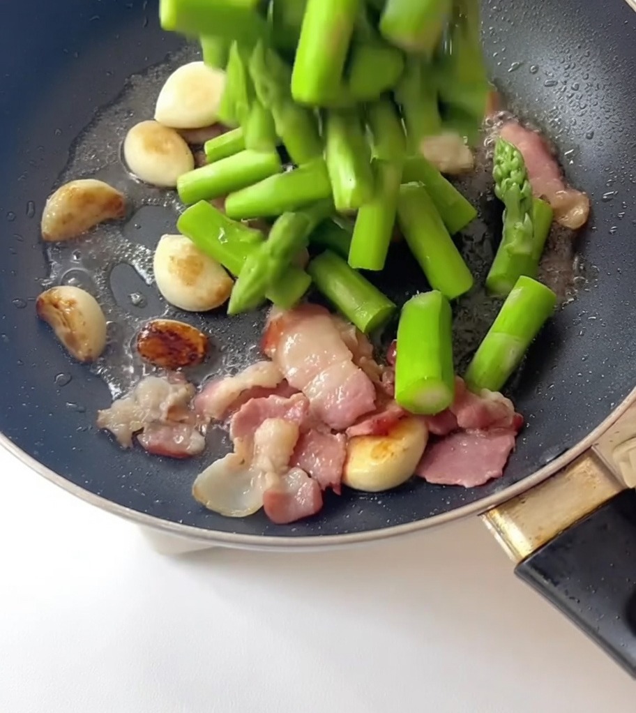 Blanched asparagus pieces being poured from a utensil into a skillet containing fried garlic cloves and bacon slices.