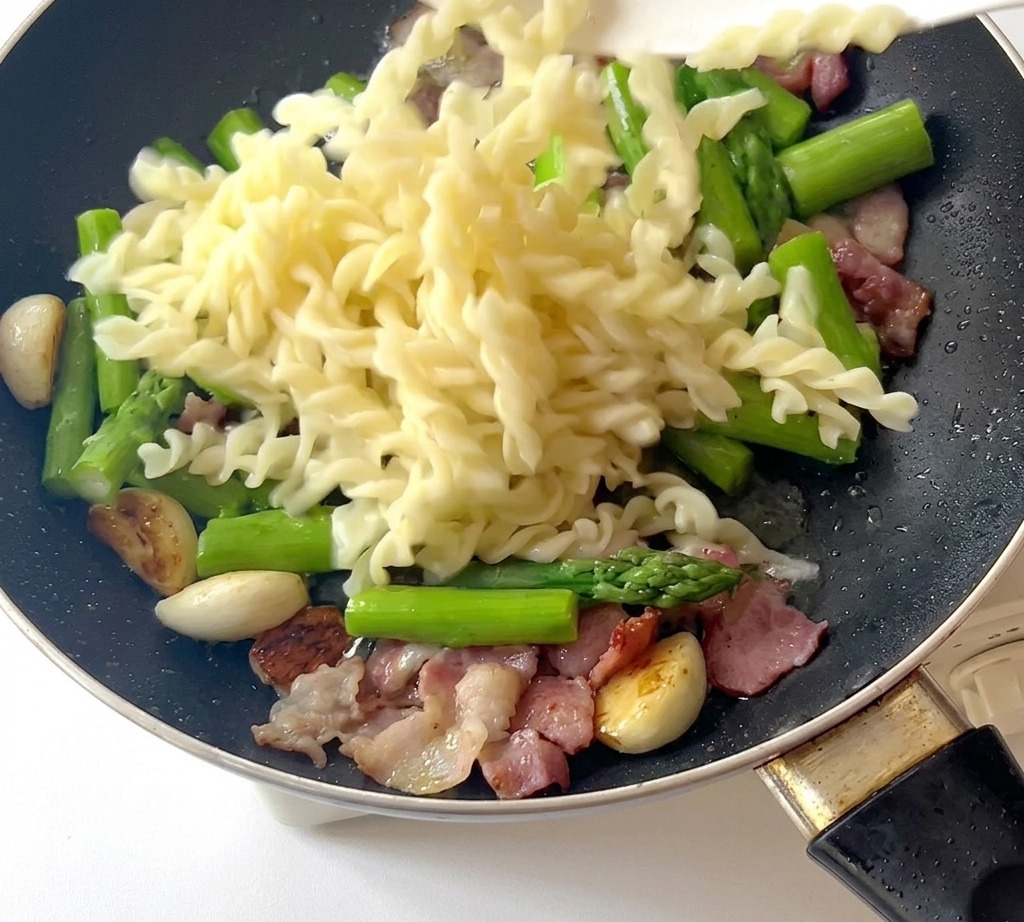 Cooked fusilli pasta being dropped into a pan already containing asparagus, bacon, and fried garlic.