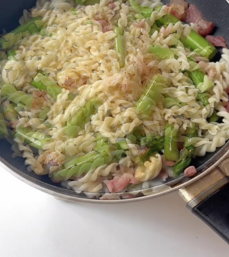 Seasonings, including black pepper, being sprinkled over a mixture of pasta, asparagus, and bacon in a frying pan.