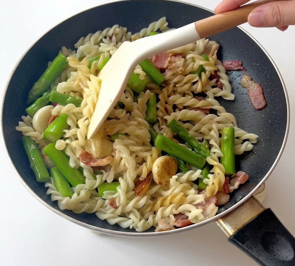 A white spatula tossing a mixture of fusilli pasta, bright green asparagus, and bacon pieces in a black skillet.