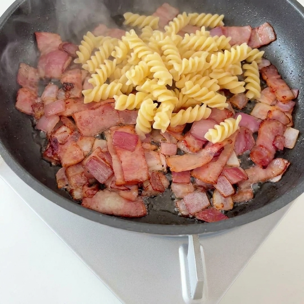 Fusilli pasta and bright green asparagus pieces sitting on top of a base of crispy bacon and caramelized onions in a frying pan.