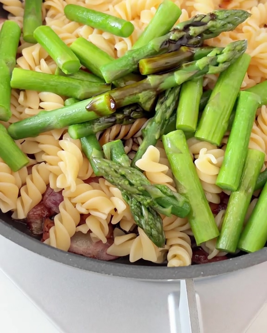Soy and oyster sauce being drizzled over a pan full of pasta, bacon, and asparagus.
