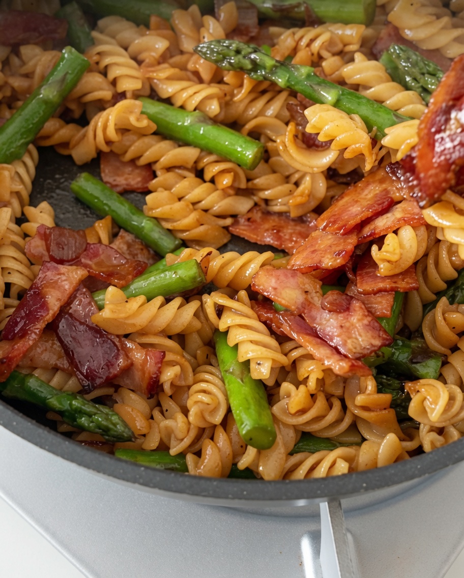 A pan of perfectly cooked fusilli pasta, crispy bacon, and tender asparagus being tossed with a glossy savory sauce.