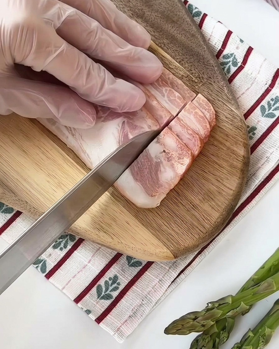Slices of raw bacon being cut into smaller, uniform squares on a wooden cutting board.