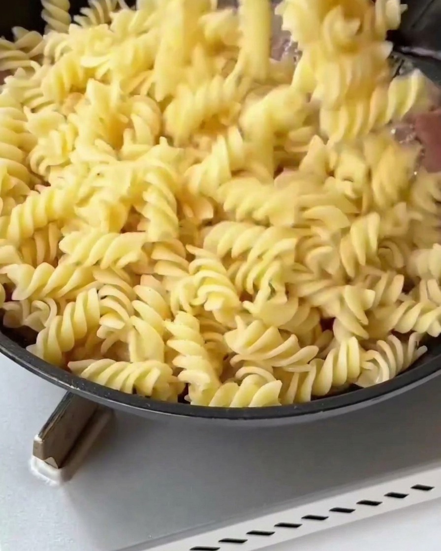 Cooked fusilli pasta and blanched asparagus pieces being added to a pan with golden fried bacon and onions.