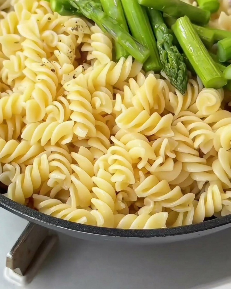 Fusilli pasta and green asparagus in a pan being seasoned with cracked black pepper.