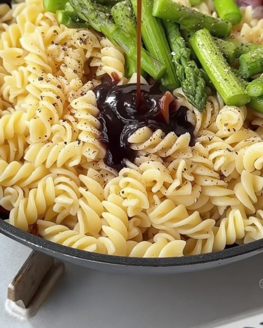 A dark, savory sauce mixture being poured from a spoon onto pasta and asparagus in a cooking pan.