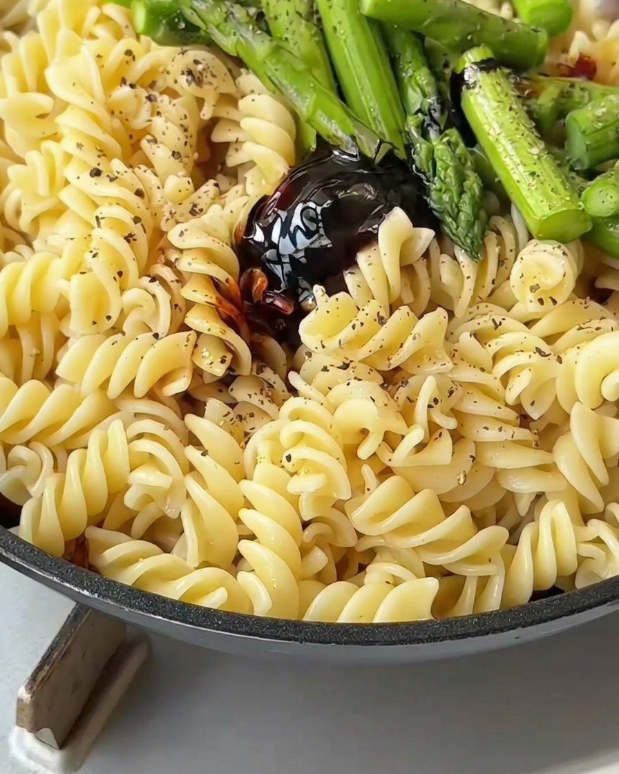 Pasta, bacon, and asparagus being tossed with a spatula in a pan to coat in savory sauce.